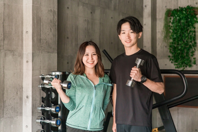 ジムで、トレーナーとお客様がダンベルを持って笑顔で並んでいる様子